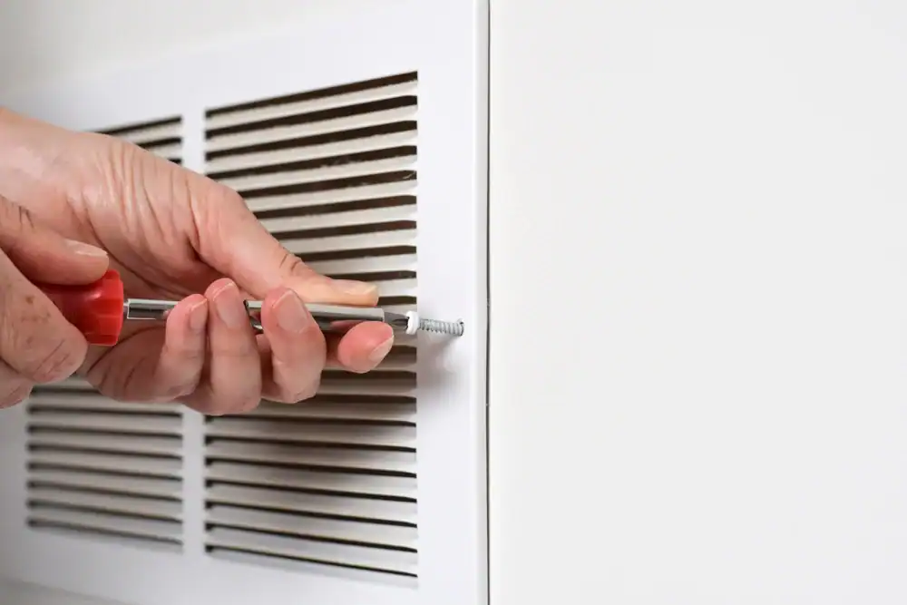 A person removes a screw from a white vent cover using a screwdriver during air duct cleaning.